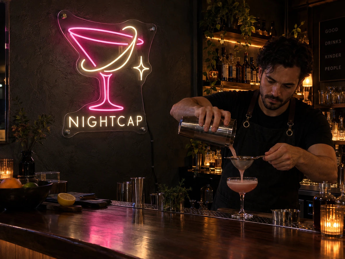 Pink and warm-white NIGHTCAP cocktail-glass neon sign in a dark bar, with a bartender preparing a drink.